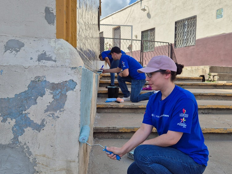 Three people in Ford blue shirts paint the lower walls of a building’s exterior steps using rollers and brushes during a community service activity.